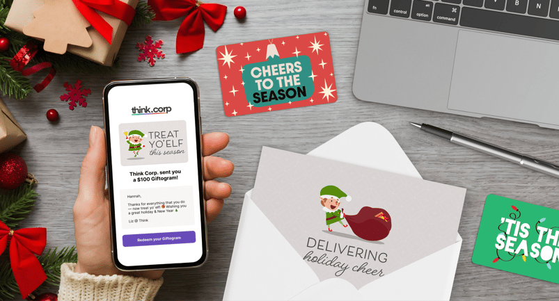 A person holds a phone showing a holiday Giftogram message, surrounded by festive decorations, gift cards with seasonal designs, and an envelope with a card featuring an elf delivering holiday cheer.