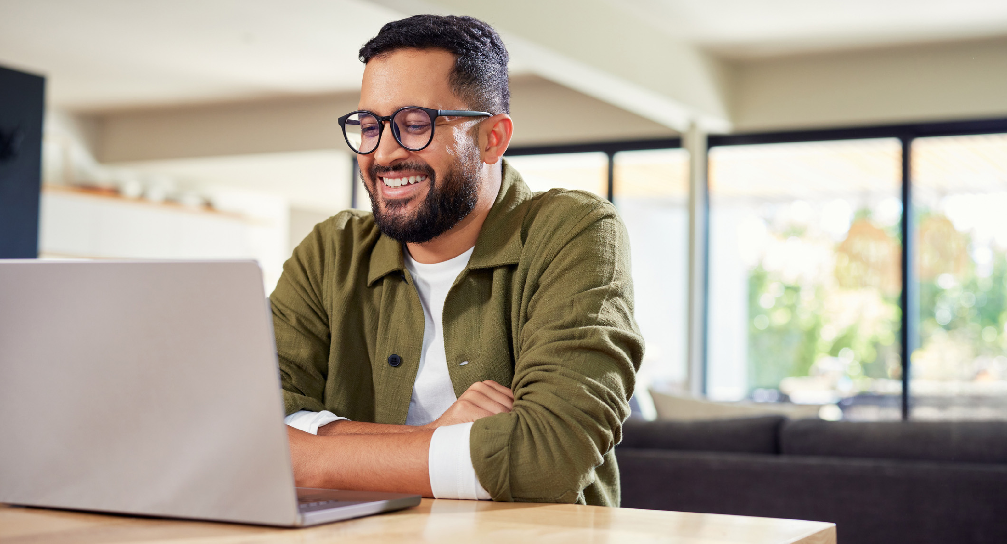Smiling man wearing glasses using a laptop at a table in a bright, modern home setting.