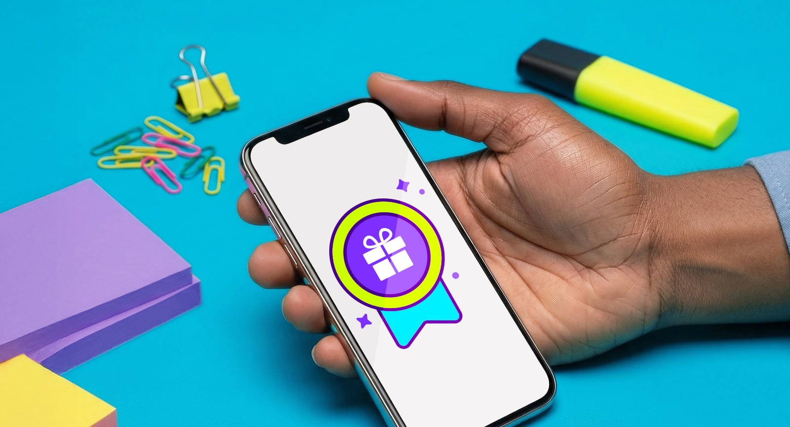 Hand holding smartphone showing a Giftogram reward badge icon. Office supplies like sticky notes and clips are on a blue desk.