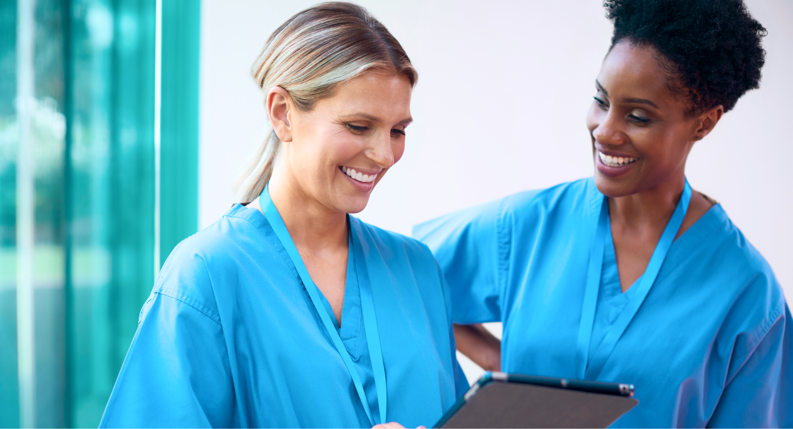 Blonde healthcare worker smiles at a tablet while a colleague watches, using Giftogram for appreciation.
