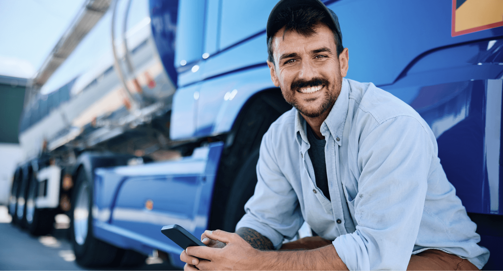 Smiling truck driver holding a smartphone, beside his blue semi-truck. Represents recognizing transportation employees for their work.