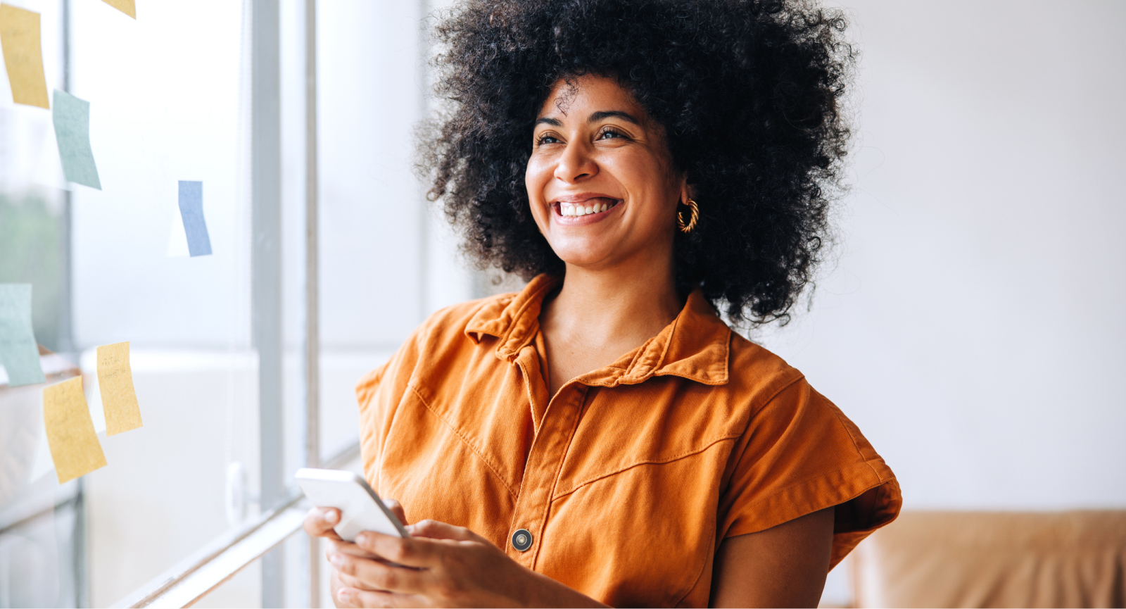 Smiling woman with afro hair, holding a phone, embodying employee happiness and recognition in a vibrant office.