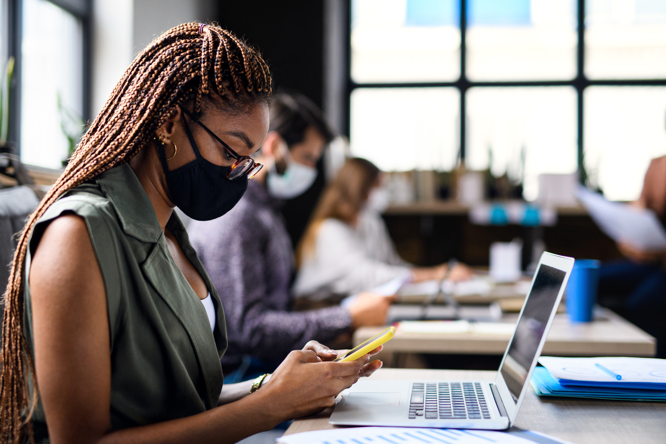 Woman with braids, glasses, and mask uses her phone at a busy office desk, representing a potential Giftogram user.