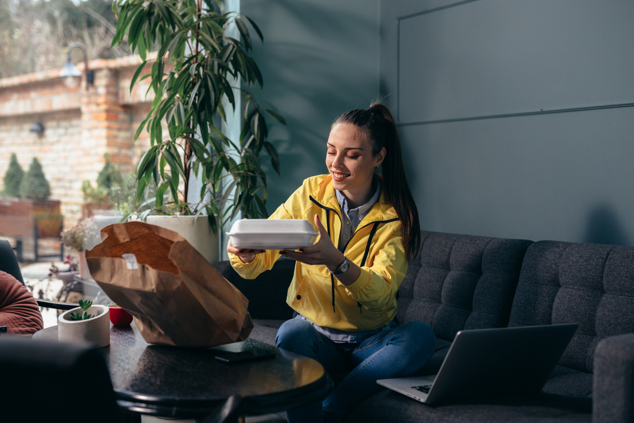 Smiling woman on a sofa opens a takeout container from a bag, enjoying a gifted meal as a token of appreciation.