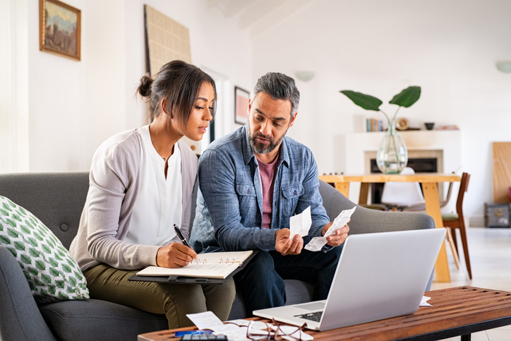 Two people on a sofa review receipts and write notes, laptop nearby, signifying financial planning or managing rewards.