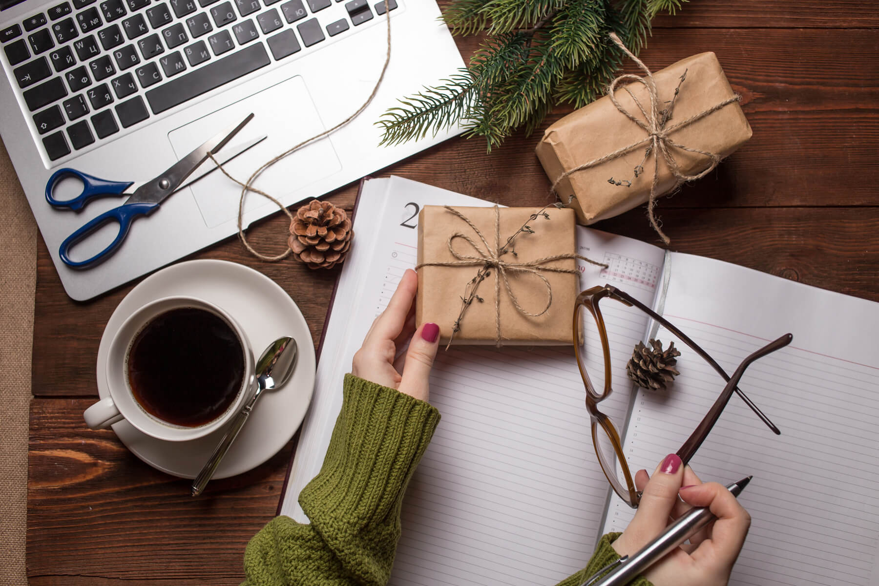 Hands hold a twine-wrapped gift on a wooden desk with a laptop and planner, planning for holiday employee appreciation.