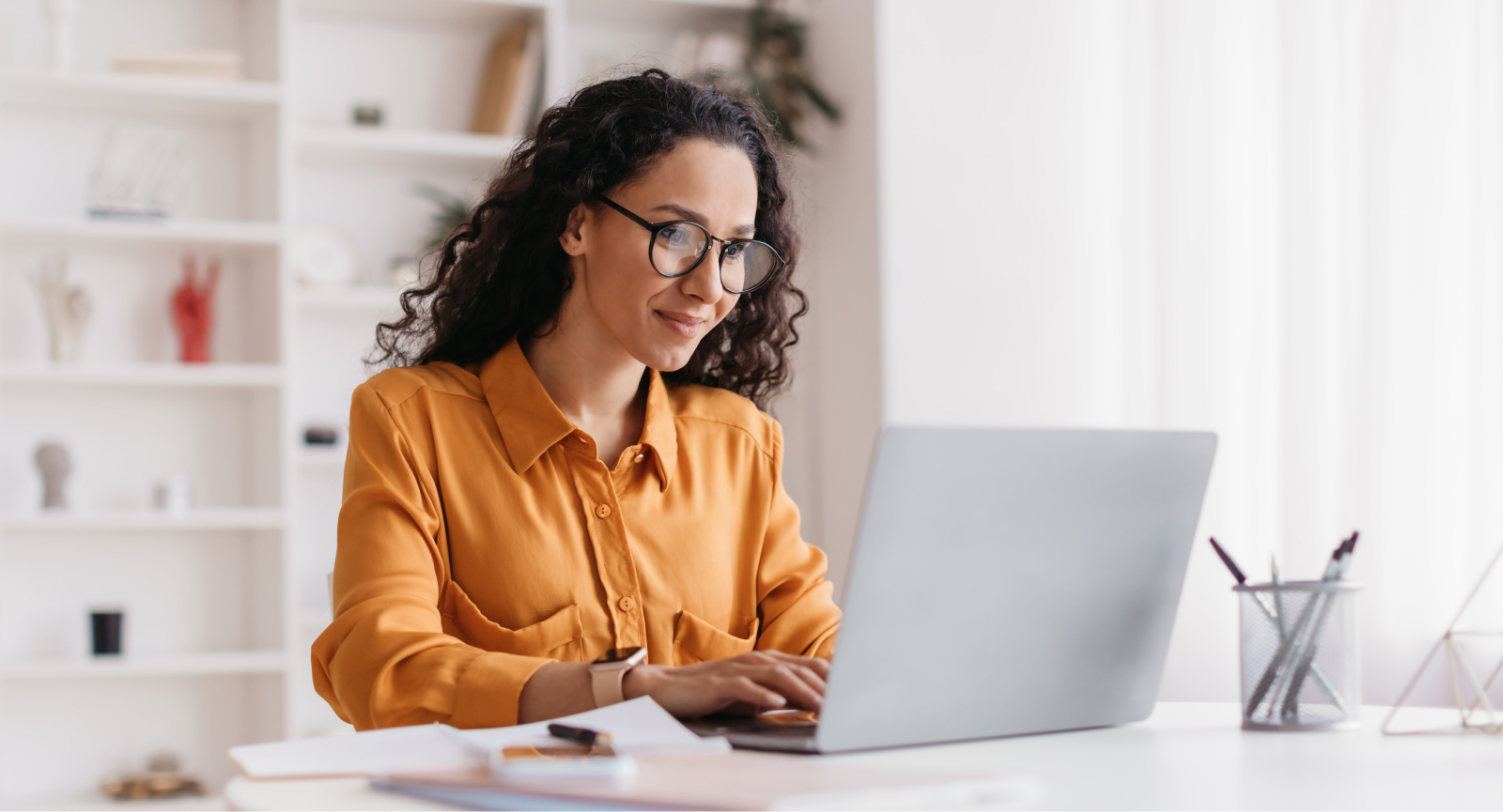 A smiling woman with curly hair and glasses types on a laptop, using Giftogram to improve employee recognition.