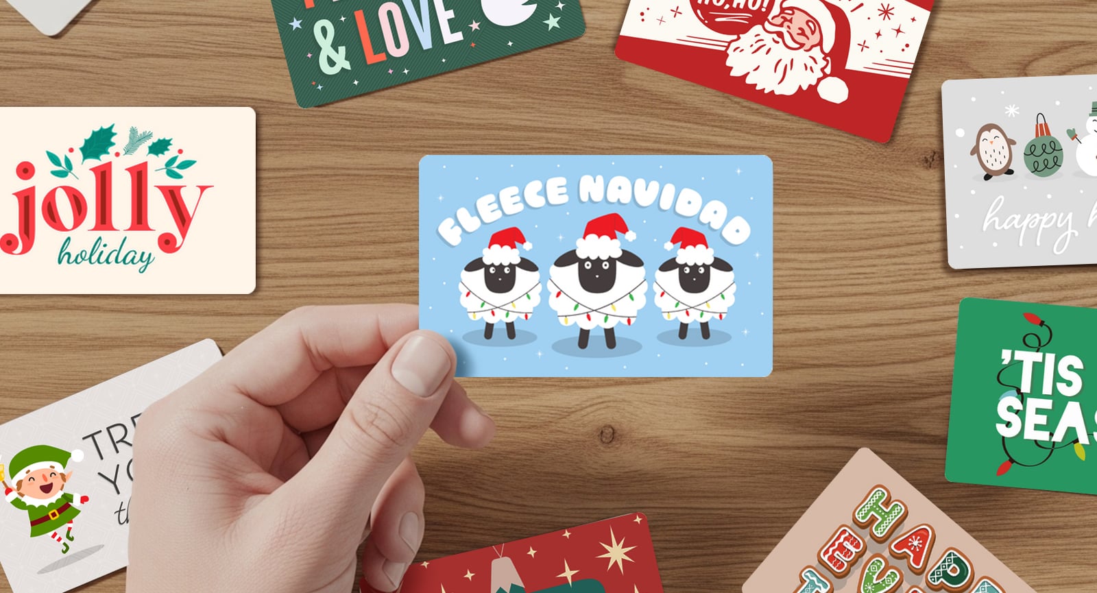 Hand holding a "FLEECE NAVIDAD" Giftogram card with three sheep, surrounded by other festive holiday gift cards on a wooden table.