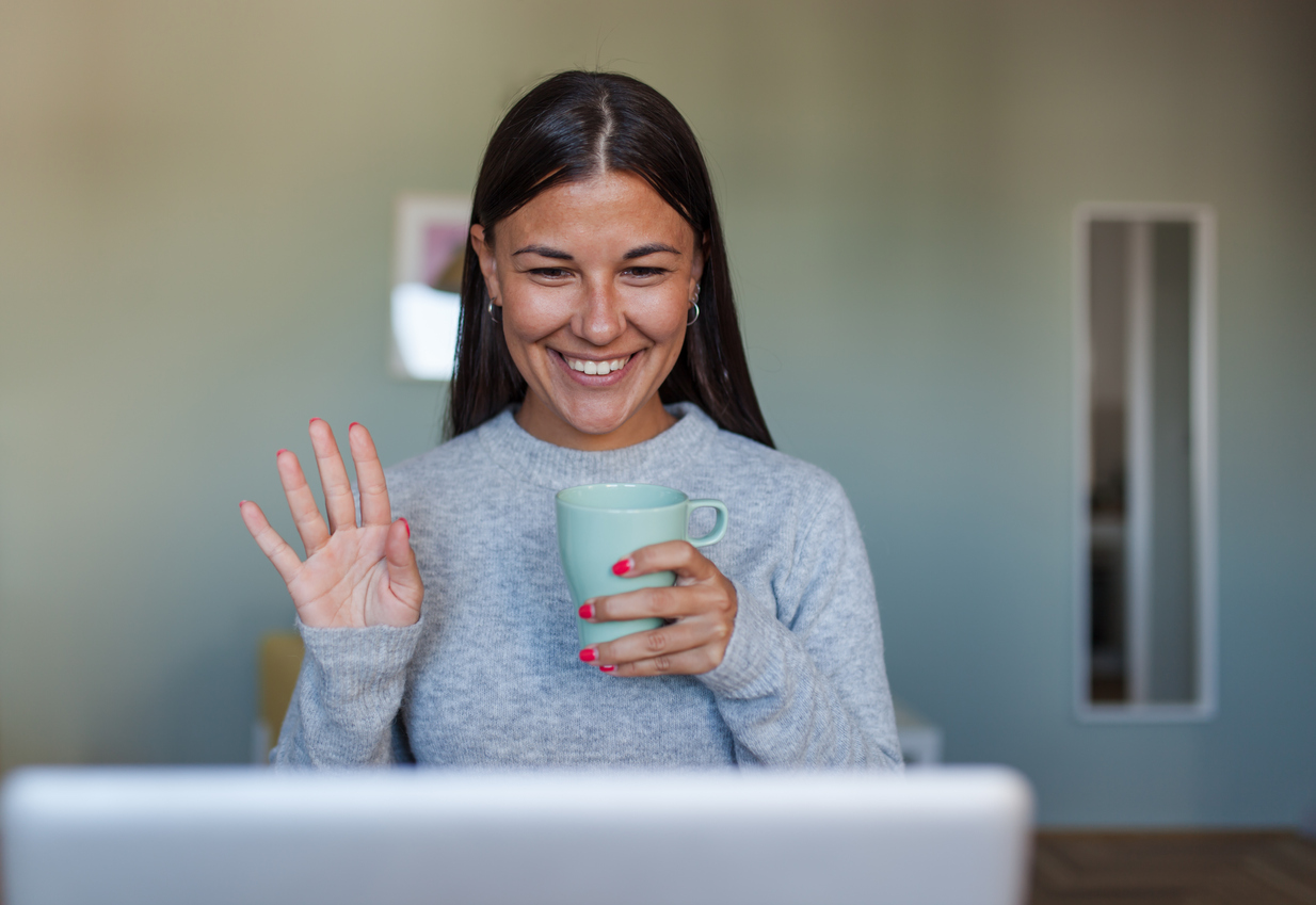 Smiling woman waving at her laptop screen, holding a mug, happily appreciating recognition.