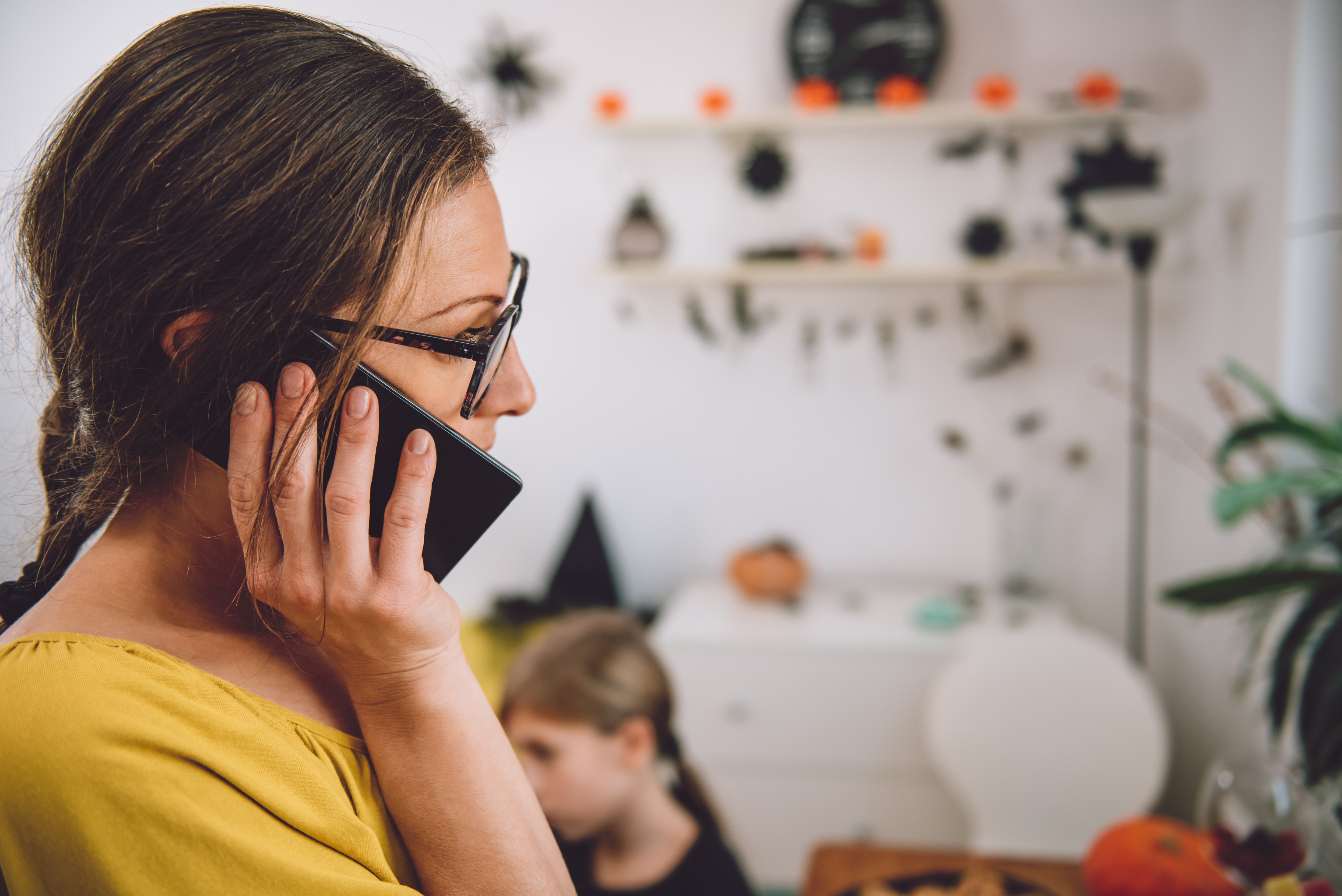 Woman in a yellow shirt and glasses talks on a phone call, in a home decorated for Halloween.